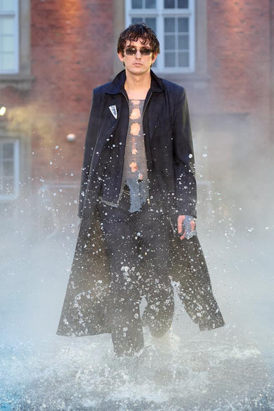 Look 14 from Deadwood FW25 Runway Show at Copenhagen Fashion Week. Model walking on a runway in the water at Villa CPH wearing navy pants and black coat in upcycled leather and Tretorn rubber boots.