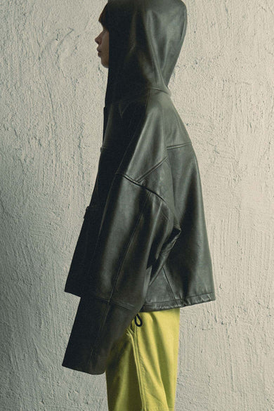 Model wearing a leather hoodie made from upcycled lambskin in forest green shot in profile. It features duo front pockets, zipper closure, a cozy hood, and a drawstring at the bottom hem.
